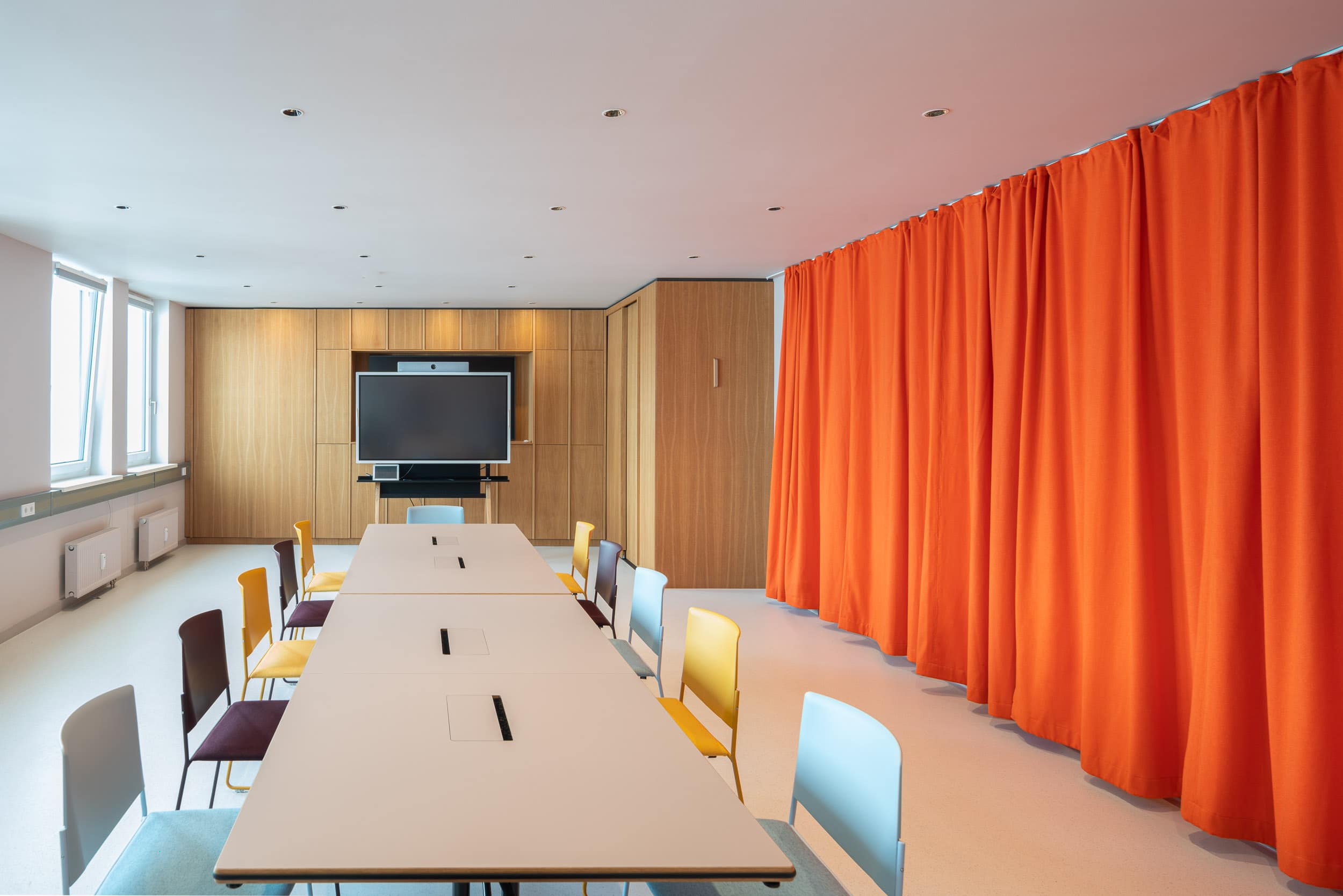 Akustikvorhang in strahlendem Orange zusammen mit farbigen Stühlen im Konferenzraum. Regalwand mit Echtholzoberfläche als individuell gestaltete Stauraumlösung.